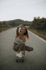 Pieno corpo attraente spensierato pattinatore femminile seduto su fianchi sullo skateboard e prendendo selfie mentre pattina sulla strada rurale vuota al crepuscolo — Foto stock