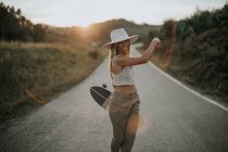 Contenuto giovane donna in abbigliamento casual e cappello estivo con in mano skateboard incrociatore e guardando la fotocamera mentre in piedi su strada asfaltata vuota nella zona rurale al tramonto — Foto stock