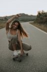 Повне тіло привабливий безтурботний жіночий ковзаняр сидить на гастролях на скейтборді і дивиться на камеру під час катання на порожній сільській дорозі в сутінках — стокове фото