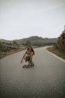 Ganzkörperattraktive unbeschwerte Skaterin sitzt auf einem Skateboard und blickt in die Kamera, während sie in der Dämmerung auf einer leeren Landstraße skatet — Stockfoto