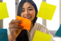 Junge fröhliche ethnische Unternehmerin arrangiert tagsüber bunte Papieraufkleber auf transparenter Oberfläche im Büro — Stockfoto