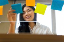 Jovem empresária étnica feliz organizando adesivos de papel coloridos na superfície transparente no escritório durante o dia — Fotografia de Stock