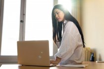 Ethnische Unternehmerin blättert im Netbook, während sie mit Papierdokument sitzt — Stockfoto
