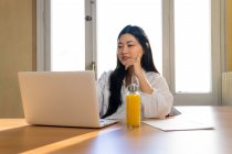 Empresaria étnica hojeando netbook mientras está sentada con documento de papel y botella de vidrio ecológico de jugo fresco - foto de stock