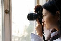 Вид збоку на урожай молода етнічна жінка фотографується на професійній цифровій камері, дивлячись вперед біля вікна в будинку — стокове фото