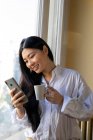 Joven mujer étnica alegre con taza de bebida caliente navegar por Internet en el teléfono celular en casa a la luz del día - foto de stock