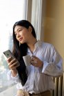 Junge fröhliche ethnische Frau mit einer Tasse Heißgetränk surft im Internet auf dem Handy zu Hause bei Tageslicht — Stockfoto