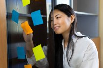 Jovem empresária étnica feliz organizando adesivos de papel coloridos na superfície transparente no escritório durante o dia — Fotografia de Stock