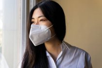 Vue de côté de la jeune femme exécutive ethnique dans un masque respiratoire regardant loin contre la fenêtre dans l'espace de travail — Photo de stock