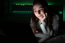 Lächelnder Teenager in lässigem Pullover liegt im Schlafzimmer, während er im dunklen Raum im Netbook blättert — Stockfoto