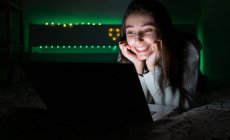 Adolescente sorridente em camisola casual deitado no quarto enquanto navega netbook no quarto escuro — Fotografia de Stock