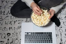From above of crop anonymous teenage in casual clothes eating popcorn while watching movie on netbook at home — Stock Photo