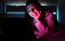 Sonriente adolescente en suéter casual acostado en el dormitorio mientras navega netbook en habitación oscura - foto de stock