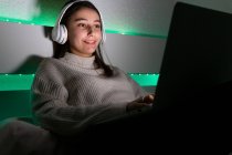 Mulher encantadora em suéter ouvir música com fones de ouvido enquanto navega laptop na cama — Fotografia de Stock