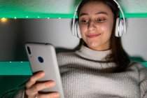 Cortar encantadora fêmea em suéter ouvir música com fones de ouvido enquanto navega telefone celular na cama — Fotografia de Stock