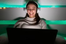 Charmante Frau im Pullover hört Musik mit Kopfhörern, während sie auf dem Bett im Laptop surft — Stockfoto