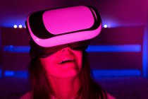 Молода темноволоса жінка в окулярах VR, дивлячись навколо, сидячи в кімнаті з яскравим освітленням — стокове фото