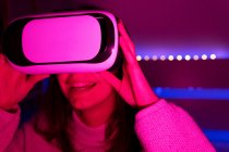 Joven hembra morena con gafas VR mirando a su alrededor mientras está sentada en la habitación con iluminación vívida - foto de stock