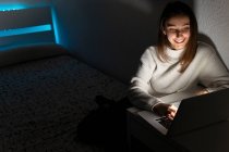 Sonriente adolescente en suéter casual sentado en el dormitorio mientras navega netbook en habitación oscura - foto de stock