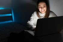Adolescent souriant en pull décontracté assis sur la chambre à coucher tout en naviguant netbook dans la chambre noire — Photo de stock