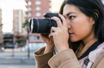 Side view of ethnic Asian female photographer shooting photo on professional photo camera on city street — Stock Photo