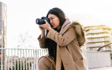 Side view of ethnic Asian female photographer shooting photo on professional photo camera on city street — Stock Photo