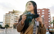 Ethnic young happy Asian female photographer shooting photo on professional photo camera on city street — Stock Photo
