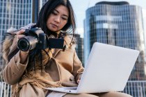 Baixo ângulo de fotógrafo feminino étnico concentrado vestindo netbook de navegação outerwear quente enquanto trabalhava remotamente — Fotografia de Stock
