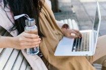 Анонімна жінка, що тримає екологічно чисту скляну пляшку з водою, працюючи дистанційно на ноутбуці, сидить на лавці на вулиці — стокове фото