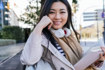 Позитивна етнічна жінка в бездротових навушниках широко посміхається, читаючи книгу, сидячи в парку і дивлячись на камеру — стокове фото