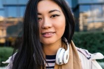 Усміхнена етнічна жінка в пальто з навушниками навколо шиї стоїть на тлі сучасної будівлі — стокове фото