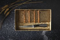 Vista superior de pan de centeno casero fresco cerca de cuchillo en canasta de mimbre y espigas de trigo en la mesa - foto de stock