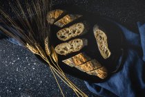 Von oben appetitlich knuspriges Brot in der Nähe von Weizenspitzen und dunklem Stoff auf dem Tisch — Stockfoto