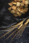 Кусочки хрустящего хлеба в миске рядом с пшеничными шипами на черном текстиле в комнате — стоковое фото