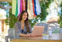 Young content female in ornamental clothes looking at camera while sitting at desk with netbook and cup of hot drink — Stock Photo