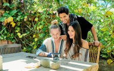 Young glad multiethnic partners taking self portrait on photo camera at table against climbing plants in courtyard — Stock Photo