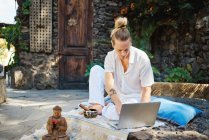 Young male surfing internet on netbook while sitting against Buddha statuette and bowl gong in patio — Stock Photo