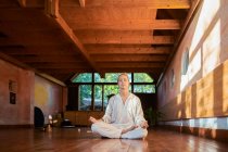 Joven hombre consciente descalzo macho sentado en pose de loto con los ojos cerrados practicando yoga en el suelo cerca del gong bowl y la estatuilla de Buda - foto de stock