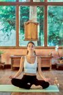 Jovem fêmea concentrada com olhos fechados praticando ioga com pernas cruzadas perto de tigela gongo em casa — Fotografia de Stock