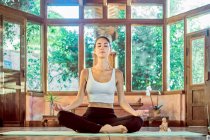 Joven hembra concentrada con los ojos cerrados practicando yoga con las piernas cruzadas cerca del bowl gong en casa - foto de stock