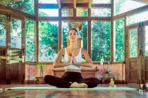Jeune femme concentrée avec les yeux fermés pratiquant le yoga avec les jambes croisées près gong bol dans la maison — Photo de stock