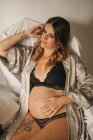 Calm pregnant female in underwear sitting with closed eyes and touching belly in light room in apartment — Stock Photo