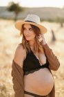 Allegro donna incinta indossa un cappello lingerie e cardigan in piedi tra erba secca in campo collocato in campagna e guardando lontano nella giornata di sole — Foto stock