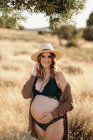 Fröhliche schwangere Frau mit Hut und Strickjacke steht zwischen trockenem Gras auf einem Feld im Grünen und blickt bei sonnigem Wetter in die Kamera — Stockfoto
