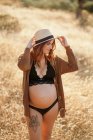 Mujer embarazada pensativa con un sombrero de lencería y cárdigan de pie entre la hierba seca en el campo colocado en el campo y mirando hacia abajo en el día soleado - foto de stock