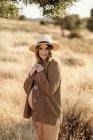 Mujer embarazada alegre con un sombrero de lencería y cárdigan de pie entre la hierba seca en el campo colocado en el campo y mirando a la cámara en el día soleado - foto de stock