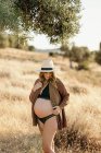 Страстная беременная женщина в шляпном белье и кардигане стоит среди сухой травы в поле, помещенной в сельской местности и глядя вниз в солнечный день — стоковое фото