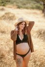 Mujer embarazada alegre con un sombrero de lencería y cárdigan de pie entre la hierba seca en el campo colocado en el campo y mirando hacia otro lado en el día soleado - foto de stock