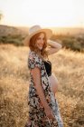 Pensiva mulher grávida em chapéu de lingerie e casaco de lã em pé entre grama seca no campo colocado no campo e olhando para longe no dia ensolarado — Fotografia de Stock