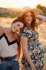 Charming female in trendy outfit leaning head on positive pregnant female while standing in field in countryside and looking at camera in sunny day — Stock Photo